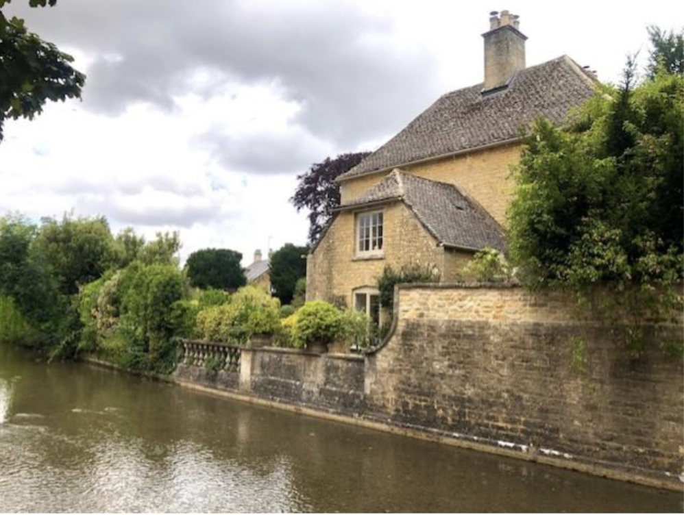 a river runs up against a stone wall and a yellow house in the Cotswold, England